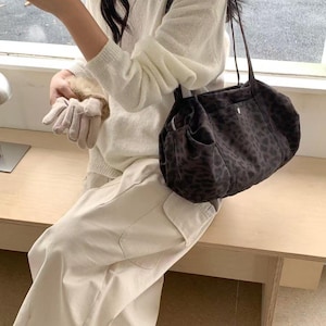 Puede incluir: Un bolso de hombro marr&oacute;n oscuro con estampado de leopardo. El bolso tiene forma redondeada y una correa larga. El bolso est&aacute; hecho de un material suave. La persona lleva un su&eacute;ter blanco y pantalones claros.