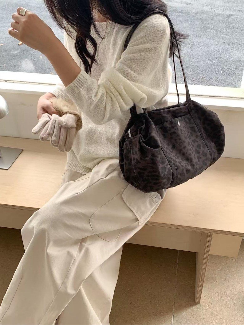 Puede incluir: Un bolso de hombro marr&oacute;n oscuro con estampado de leopardo. El bolso tiene forma redondeada y una correa larga. El bolso est&aacute; hecho de un material suave. La persona lleva un su&eacute;ter blanco y pantalones claros.