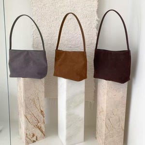 May include: Three suede shoulder bags in gray, brown, and burgundy are displayed on marble pedestals. Each bag has a slender handle and a rectangular shape. The bags are presented against a neutral background, highlighting their colors and textures.