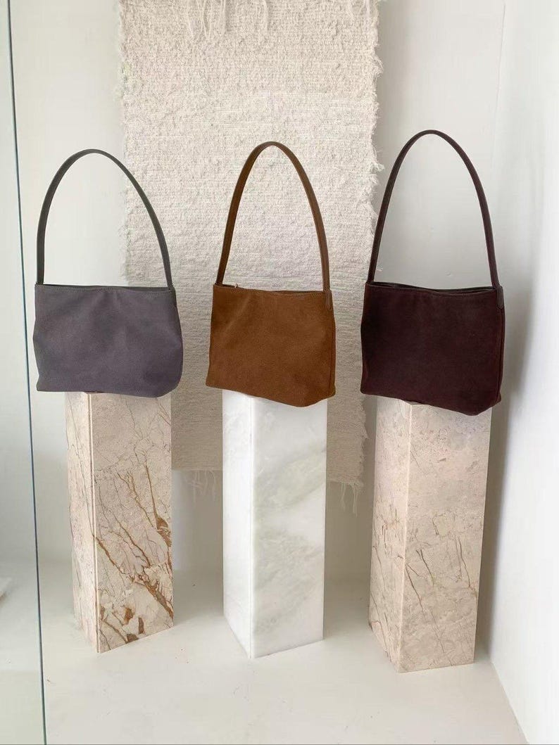 May include: Three suede shoulder bags in gray, brown, and burgundy are displayed on marble pedestals. Each bag has a slender handle and a rectangular shape. The bags are presented against a neutral background, highlighting their colors and textures.