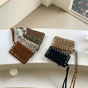 May include: An assortment of small, rectangular coin purses in various colors and patterns. The purses include brown, black, and leopard print designs, some with silver stud accents and wrist straps.
