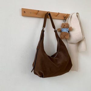 May include: A brown leather hobo bag hangs from a wooden peg rack. The bag has a curved shape and a long strap. A small teddy bear keychain and a cream-colored scarf are also hanging on the rack.