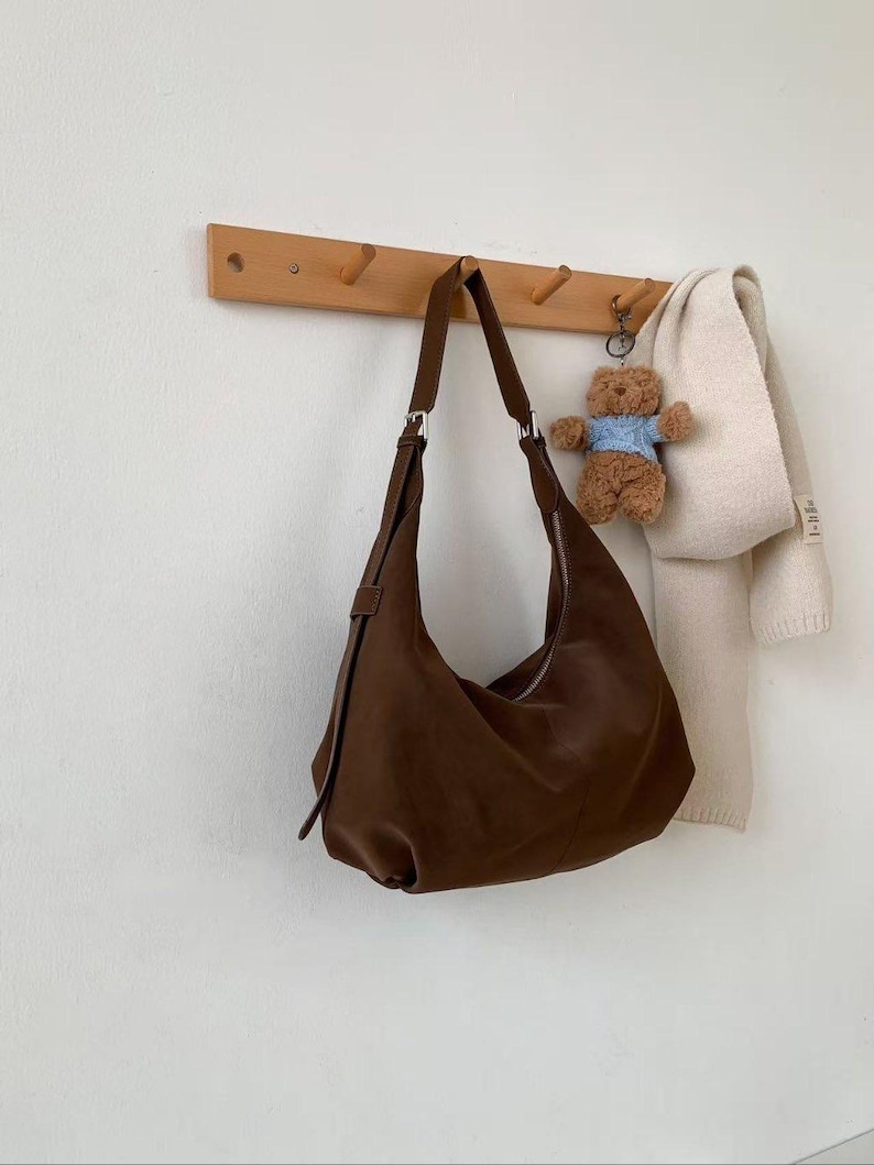 May include: A brown leather hobo bag hangs from a wooden peg rack. The bag has a curved shape and a long strap. A small teddy bear keychain and a cream-colored scarf are also hanging on the rack.