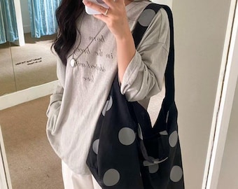 Puede incluir: Un bolso negro con lunares grises grandes. El bolso se lleva al hombro. La persona de la imagen lleva una camiseta gris de manga larga y pantalones blancos. La camiseta tiene texto.
