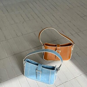 May include: Two shoulder bags. One is light blue with white trim and a silver buckle. The other is brown with white trim and a gold buckle. Both bags have a rectangular shape and a single handle.