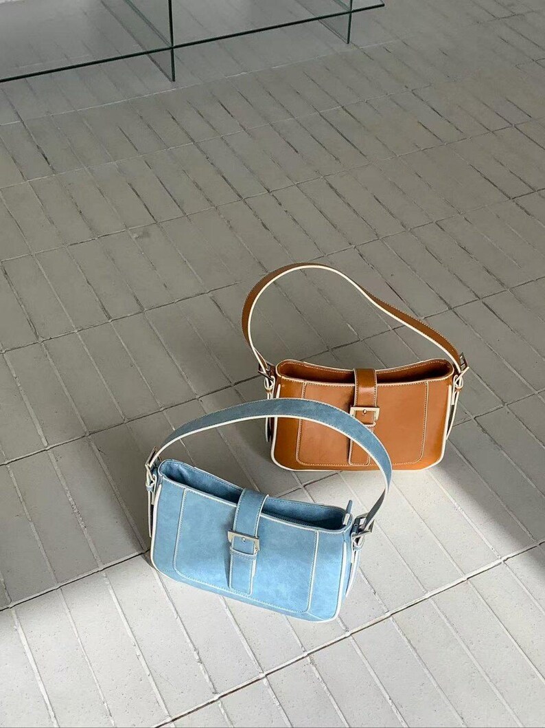 May include: Two shoulder bags. One is light blue with white trim and a silver buckle. The other is brown with white trim and a gold buckle. Both bags have a rectangular shape and a single handle.