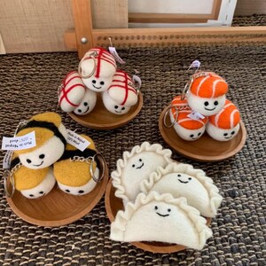 May include: Handmade felt keyrings featuring food-themed designs. The collection includes sushi, dumplings, and bees, all with smiling faces. Each set is presented on a small wooden plate.