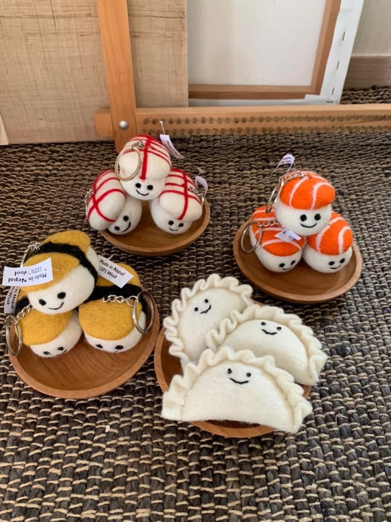 May include: Handmade felt keyrings featuring food-themed designs. The collection includes sushi, dumplings, and bees, all with smiling faces. Each set is presented on a small wooden plate.