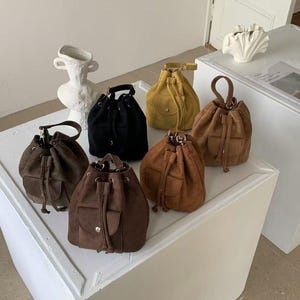 Op de afbeelding: Een collectie suède emmermodellen met trekkoord in verschillende kleuren, waaronder zwart, bruin en geel. Elke tas heeft een klein voorvak en een handvat of schouderriem. De tassen worden op een wit oppervlak gepresenteerd.