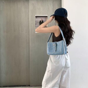May include: A light blue denim shoulder bag with a silver buckle and white trim. The bag is rectangular with a flap closure. The person is wearing a navy blue baseball cap, a brown vest top, and white trousers.