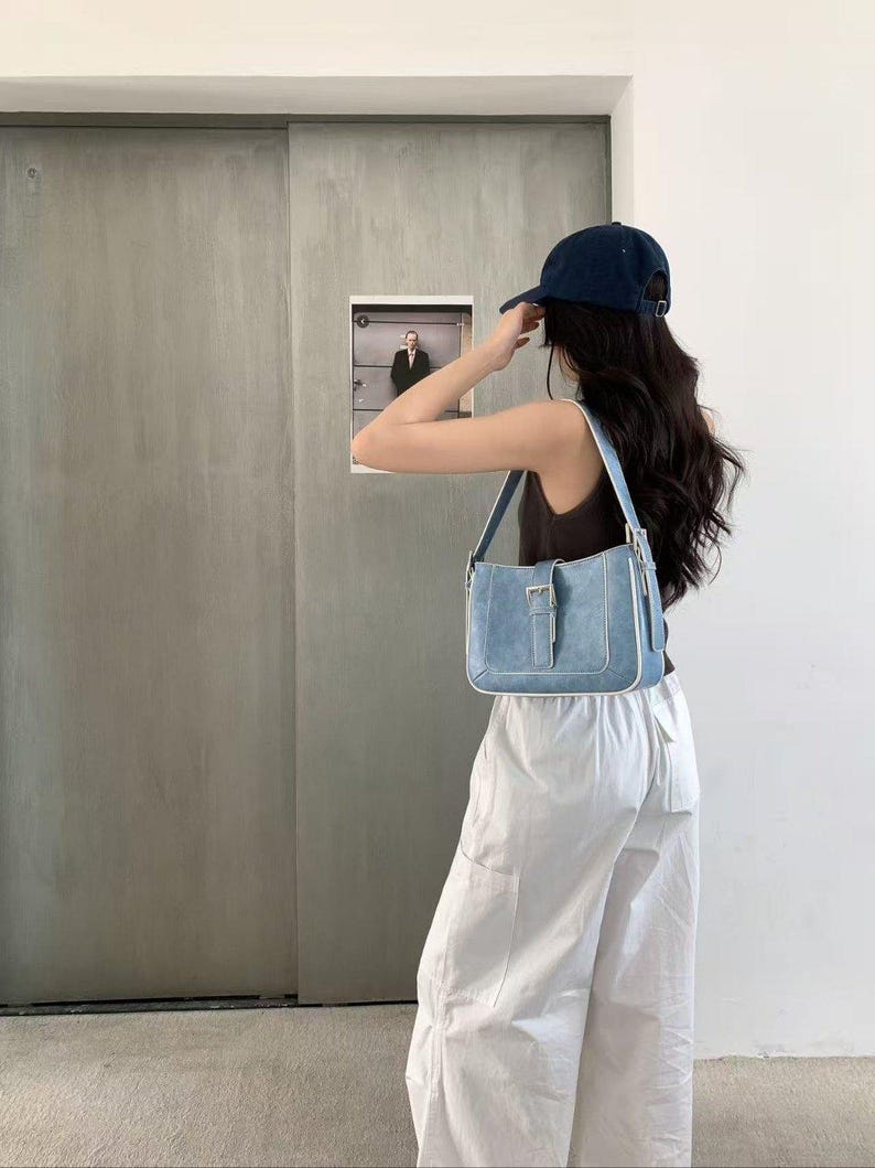 May include: A light blue denim shoulder bag with a silver buckle and white trim. The bag is rectangular with a flap closure. The person is wearing a navy blue baseball cap, a brown vest top, and white trousers.