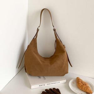 May include: A brown suede shoulder bag with a long strap. The bag is sitting on a white surface next to a book, a plate with a croissant, and a pair of brown gloves.