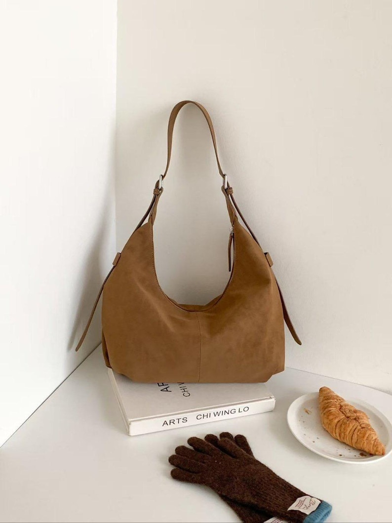 May include: A brown suede shoulder bag with a long strap. The bag is sitting on a white surface next to a book, a plate with a croissant, and a pair of brown gloves.