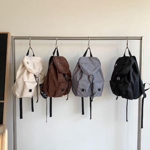 May include: Four backpacks in cream, brown, gray, and black hang from a metal rack. Each backpack has a drawstring closure, a front pocket, and black straps. The backpacks are made of a durable material and are suitable for everyday use.