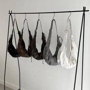 Pode incluir: Exibição de cinco bolsas de ombro em várias cores: preto, castanho, castanho escuro, cinzento e branco. As bolsas estão penduradas num cabide de metal preto contra uma parede branca. Cada bolsa tem uma alça comprida.