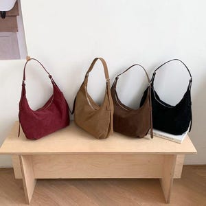 May include: Four suede shoulder bags in shades of burgundy, tan, brown, and black are displayed on a light wood table. Each bag has a curved shape, a zipper closure, and a single shoulder strap. The bags are arranged in a row, showcasing their different colors.