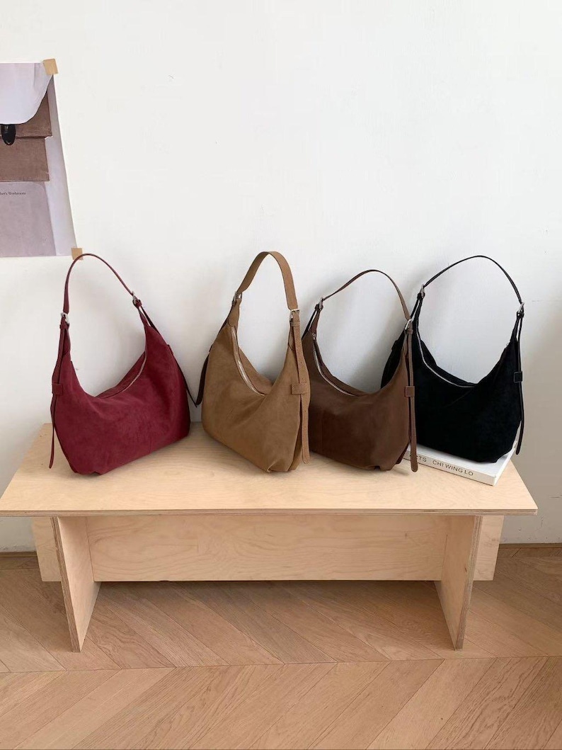 May include: Four suede shoulder bags in shades of burgundy, tan, brown, and black are displayed on a light wood table. Each bag has a curved shape, a zipper closure, and a single shoulder strap. The bags are arranged in a row, showcasing their different colors.
