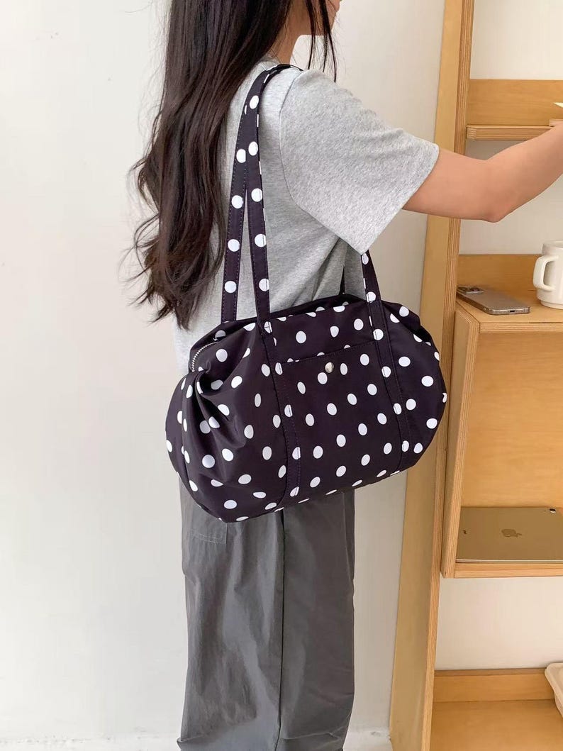 Puede incluir: Bolso tote negro con lunares blancos, con cierre de cremallera y dos asas. El bolso cil&iacute;ndrico tiene un peque&ntilde;o bolsillo delantero. El material es suave y flexible.