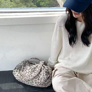 Puede incluir: Bolso tote con estampado de leopardo en tonos grises y crema, con dos asas. Tambi&eacute;n se ve una gorra azul marino con la palabra "Amptons". El bolso est&aacute; sobre una superficie oscura.