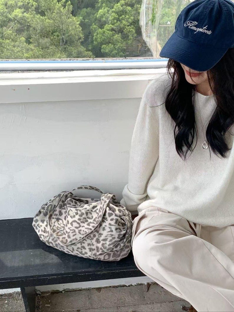 Puede incluir: Bolso tote con estampado de leopardo en tonos grises y crema, con dos asas. Tambi&eacute;n se ve una gorra azul marino con la palabra "Amptons". El bolso est&aacute; sobre una superficie oscura.
