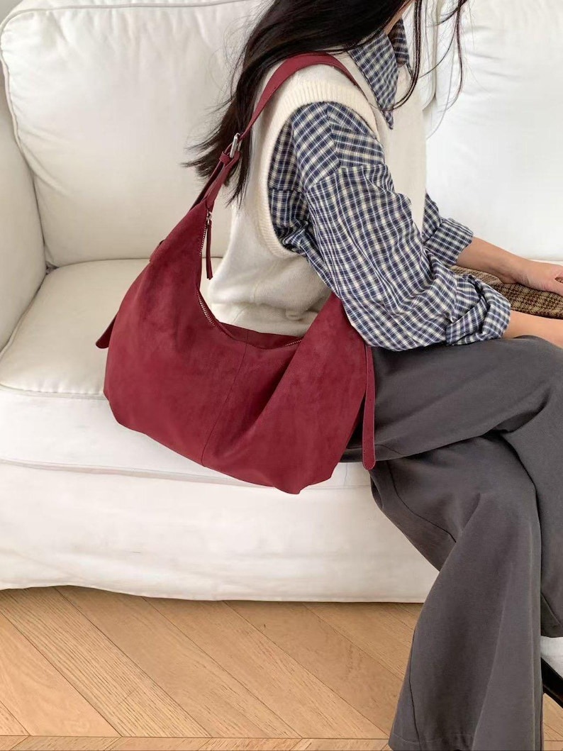 May include: A burgundy suede shoulder bag with a long strap. The bag has a curved shape and a zipper closure. The bag is a deep red color and appears to be made of soft material. The bag is a stylish accessory.