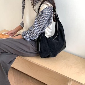 May include: A black suede shoulder bag with a silver zipper closure. The bag is worn over the shoulder. The person is wearing a white sweater vest over a blue and white plaid shirt and gray pants.
