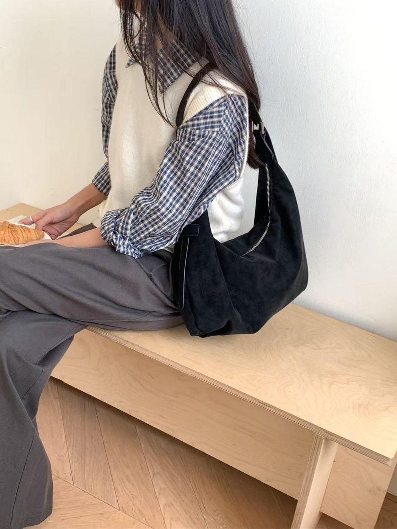 May include: A black suede shoulder bag with a silver zipper closure. The bag is worn over the shoulder. The person is wearing a white sweater vest over a blue and white plaid shirt and gray pants.