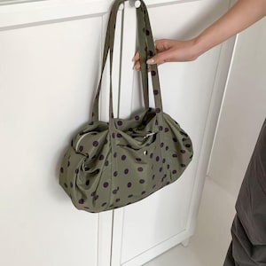 Puede incluir: Bolso tote verde oliva con lunares morados oscuros. El bolso tiene dos asas largas y una cremallera en la parte superior. Un peque&ntilde;o bolsillo es visible en la parte delantera del bolso.