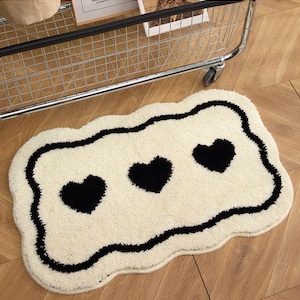Puede incluir: Una alfombra en forma de corazón de color crema con tres corazones negros y un borde negro ondulado. La alfombra está hecha de un material suave y texturizado y está colocada sobre un suelo de madera. Un artículo decorativo para un dormitorio o baño.