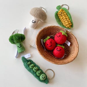 May include: A set of felt food keychains featuring a mushroom, corn on the cob, broccoli, peas in a pod, and three tomatoes. Each keychain has a smiling face.