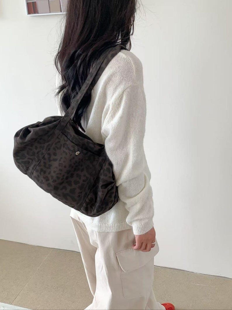 Puede incluir: Un bolso de hombro marr&oacute;n oscuro con estampado de leopardo. El bolso se lleva sobre el hombro. La persona lleva un su&eacute;ter blanco y pantalones color beige.