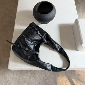 Puede incluir: Bolso de hombro de cuero negro con hebillas y correas plateadas. El bolso tiene un dise&ntilde;o asim&eacute;trico &uacute;nico. Un cuenco decorativo negro y redondo est&aacute; cerca sobre una superficie blanca.