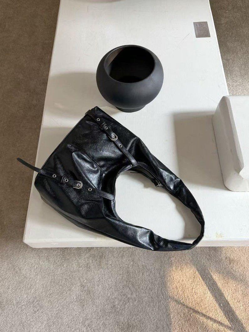Puede incluir: Bolso de hombro de cuero negro con hebillas y correas plateadas. El bolso tiene un dise&ntilde;o asim&eacute;trico &uacute;nico. Un cuenco decorativo negro y redondo est&aacute; cerca sobre una superficie blanca.