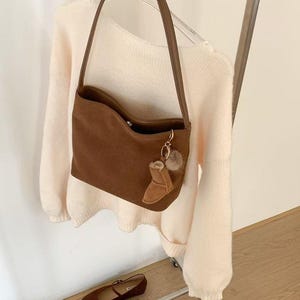 May include: A brown suede shoulder bag with a matching strap and a small boot-shaped charm. The bag is hanging on a clothing rack with a cream-colored sweater. A pair of brown flats are on a shelf below.