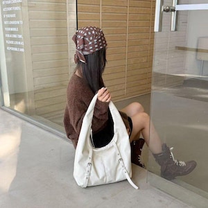 Puede incluir: Un bolso de hombro color crema con hebillas y correas plateadas. El bolso lo sostiene una persona que lleva un su&eacute;ter marr&oacute;n y un pa&ntilde;uelo en la cabeza estampado. El texto en el cristal dice: TRANSACTIONS AFFECT THE OF YOUR CARD AND ACCOUNT FUNDS IF YOU HAVE NOT LOGGED IN TO YOUR MOBILE FOR A LONG TIME PLEASE LOGIN AS AS POSSIBLE AND.