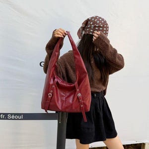 Puede incluir: Un bolso de hombro de cuero burdeos con correas ajustables y hebillas plateadas. El bolso tiene un dise&ntilde;o holgado y un asa larga y curvada. El bolso lo sostiene una persona que lleva un su&eacute;ter marr&oacute;n y una falda negra.