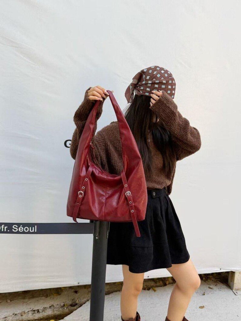 Puede incluir: Un bolso de hombro de cuero burdeos con correas ajustables y hebillas plateadas. El bolso tiene un dise&ntilde;o holgado y un asa larga y curvada. El bolso lo sostiene una persona que lleva un su&eacute;ter marr&oacute;n y una falda negra.