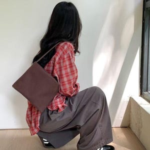 May include: A brown shoulder bag with a rectangular shape. The person is wearing a red plaid shirt and gray pants. The shoes are black with white stripes.