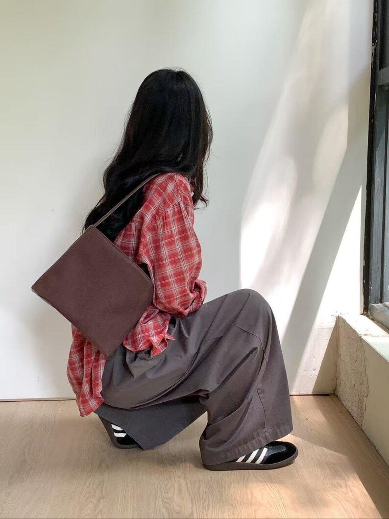 May include: A brown shoulder bag with a rectangular shape. The person is wearing a red plaid shirt and gray pants. The shoes are black with white stripes.