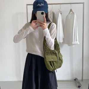 Op de afbeelding: Een marineblauwe baseballcap met witte letters, een lichtgekleurde trui en een zwarte rok. Een olijfgroene schoudertas met zakken is zichtbaar. Witte shirts met lange mouwen hangen op een kledingrek op de achtergrond.