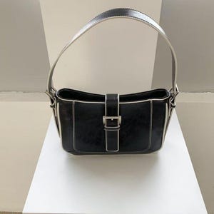 May include: Black shoulder bag with a rectangular shape, white stitching, and a silver buckle closure. The bag has a curved handle with a white interior and a black exterior. The bag is sitting on a white surface.