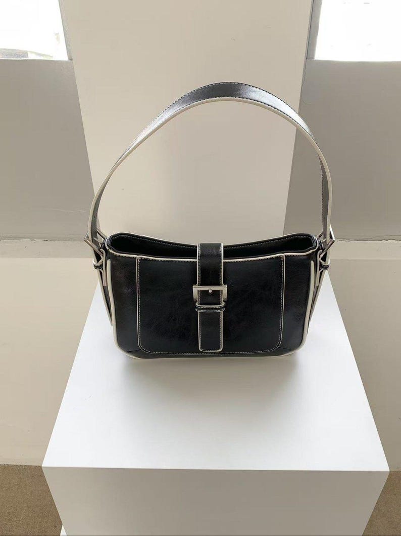 May include: Black shoulder bag with a rectangular shape, white stitching, and a silver buckle closure. The bag has a curved handle with a white interior and a black exterior. The bag is sitting on a white surface.