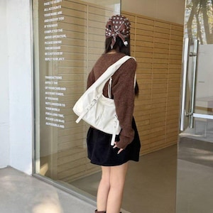Puede incluir: Una persona con un su&eacute;ter marr&oacute;n, una falda negra y botas marrones, lleva un bolso blanco. El bolso tiene hebillas plateadas y una correa larga. El texto en el cristal dice: "IN THE UPPER RIGHT CORNER OF THE PAGE TO VERIFY ALL ACCOUNTS..."