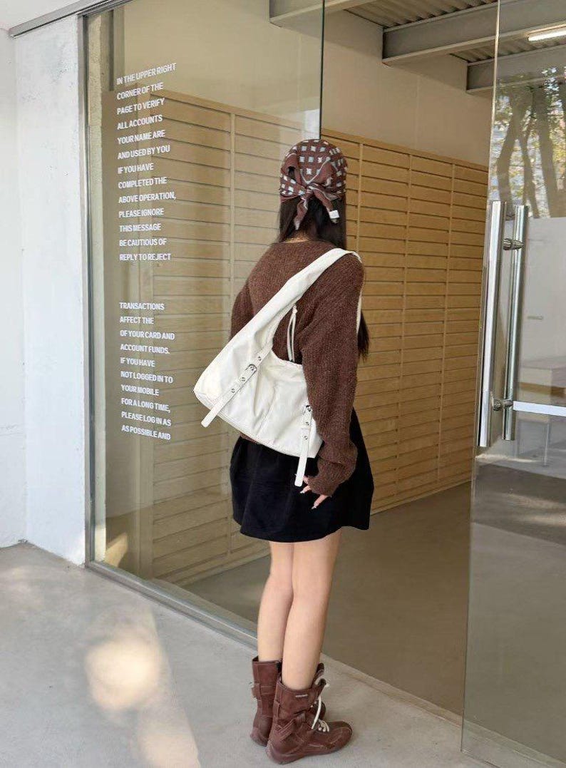 Puede incluir: Una persona con un su&eacute;ter marr&oacute;n, una falda negra y botas marrones, lleva un bolso blanco. El bolso tiene hebillas plateadas y una correa larga. El texto en el cristal dice: "IN THE UPPER RIGHT CORNER OF THE PAGE TO VERIFY ALL ACCOUNTS..."