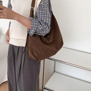 May include: A brown suede shoulder bag with a long strap. The bag is worn over a white sweater vest and a blue and white plaid shirt. The person is also wearing gray pants.