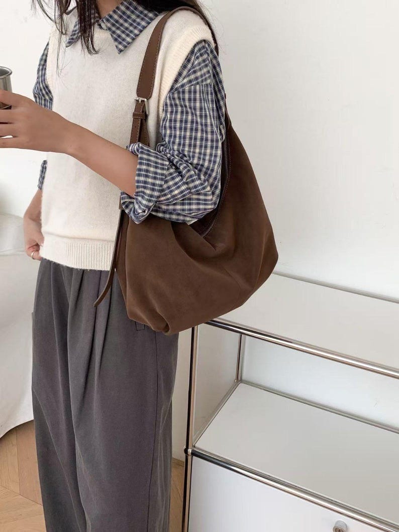May include: A brown suede shoulder bag with a long strap. The bag is worn over a white sweater vest and a blue and white plaid shirt. The person is also wearing gray pants.