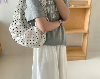 Puede incluir: Un bolso tote blanco con lunares negros y una correa con volantes. El bolso se lleva al hombro. La persona lleva una camiseta gris y pantalones blancos.