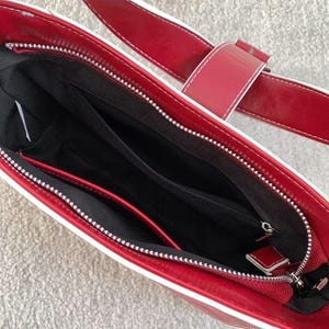 May include: A red handbag with white edging and a matching strap. The bag has a silver zip and a black interior with several compartments. The strap features a buckle fastening. The bag is made of a glossy material.