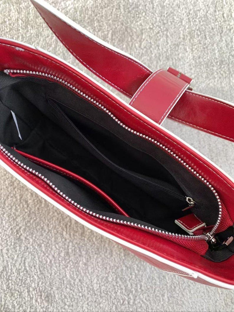 May include: A red handbag with white edging and a matching strap. The bag has a silver zip and a black interior with several compartments. The strap features a buckle fastening. The bag is made of a glossy material.