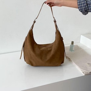 May include: A brown suede shoulder bag with a curved design. The bag has a long strap and silver-colored hardware. The bag is on a white surface. A hand is holding the strap. A small perfume bottle and a folded scarf are also on the surface.